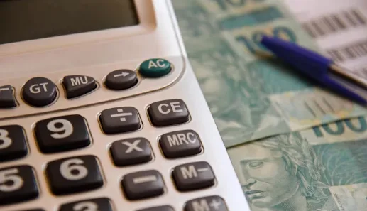 Imagem para referenciar carga tributária, reforma tributária, tributos e impostos. Calculadora, notas de 100 reais e caneta.